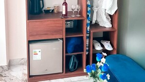 A hotel room with a wooden wardrobe, a small refrigerator, a coffee maker, wine glasses, a bottle of wine, a pair of slippers, and a vase with flowers. - Hotel BlueThree Chancay (Chancay)