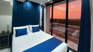 A hotel room with a large bed, a nightstand, a window with a view of the sunset, and a balcony. - Hotel BlueThree Chancay (Chancay)