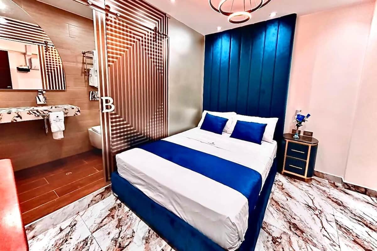 A modern bedroom with a large bed, a nightstand, a mirror, and a bathroom visible in the background.