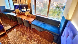 A modern dining area with a long table, blue cushioned seating, and large windows offering a view of a town and mountains. - Hotel BlueThree Chancay (Chancay)
