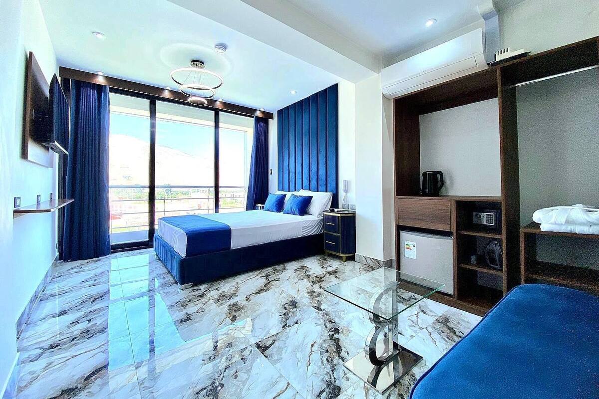 A modern hotel room with a large bed, a glass coffee table, a wardrobe, and a view of the city.