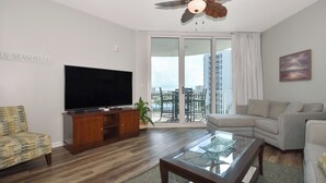 A modern living room with a flat-screen TV, wooden entertainment unit, glass coffee table, and a sofa. - 6th Floor condo at The Palms of Destin with Seasonal Beach Shuttle! (Destin)