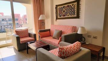 Ein Wohnzimmer mit einer Couch, zwei Sesseln, einem Couchtisch und einem Fernseher. Ein Balkon mit Tisch und Stühlen ist vorhanden.