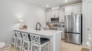 A modern kitchen with white cabinetry, a stainless steel refrigerator, a microwave, and a built-in oven. There is a white island with bar stools and a small sink.