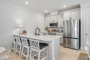 Una cocina moderna con armarios blancos, un refrigerador de acero inoxidable, un microondas y un horno empotrado. Hay una isla blanca con taburetes altos y un lavamanos pequeño.