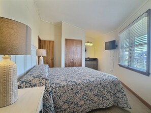 Een slaapkamer met een bed, een dressoir, een lamp, een televisie en een raam met jaloezieën.