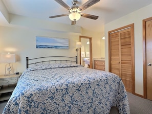 A bedroom with a bed, a ceiling fan, a nightstand, a lamp, a painting, a mirror, a wooden wardrobe, and a bathroom visible through an open door.