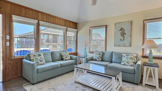 A living room with two light blue sofas, a white coffee table, and a ceiling fan. Large windows provide views of the outside. - Sea Horse- Oceanside- Only a short Walk to Beach- Community Pool & Spa (Buxton)