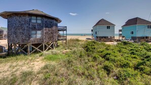 Una casa de playa con una torre de madera, dos edificios principales y una playa arenosa al fondo.