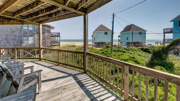 Terraza de madera con mobiliario exterior, con vista a una playa y dos casas.