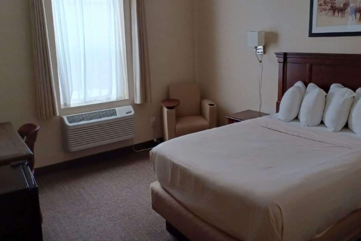 Une chambre d’hôtel avec un lit, une chaise, un bureau, une lampe et une fenêtre avec des rideaux.