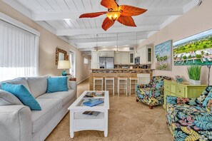 A living room with a sofa, coffee table, and a colorful armchair. There is a kitchen area with a bar and a dining table.