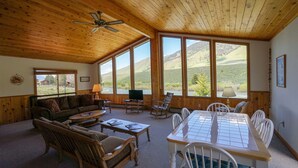 A cozy living room with wooden furniture, a dining table, and large windows offering a view of the outdoors. - Pagenkopf House on the Madison River (Cameron)