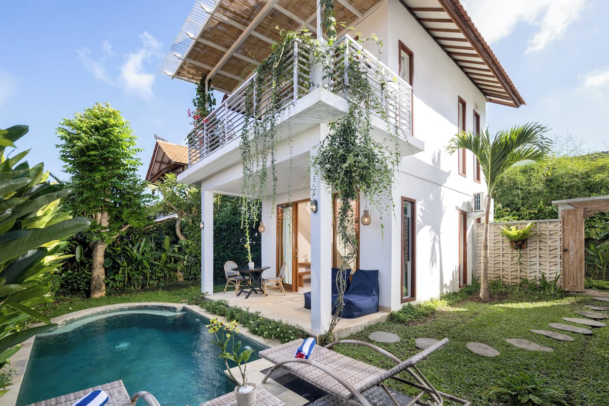 A two-story house with a balcony, a swimming pool, and lush greenery.