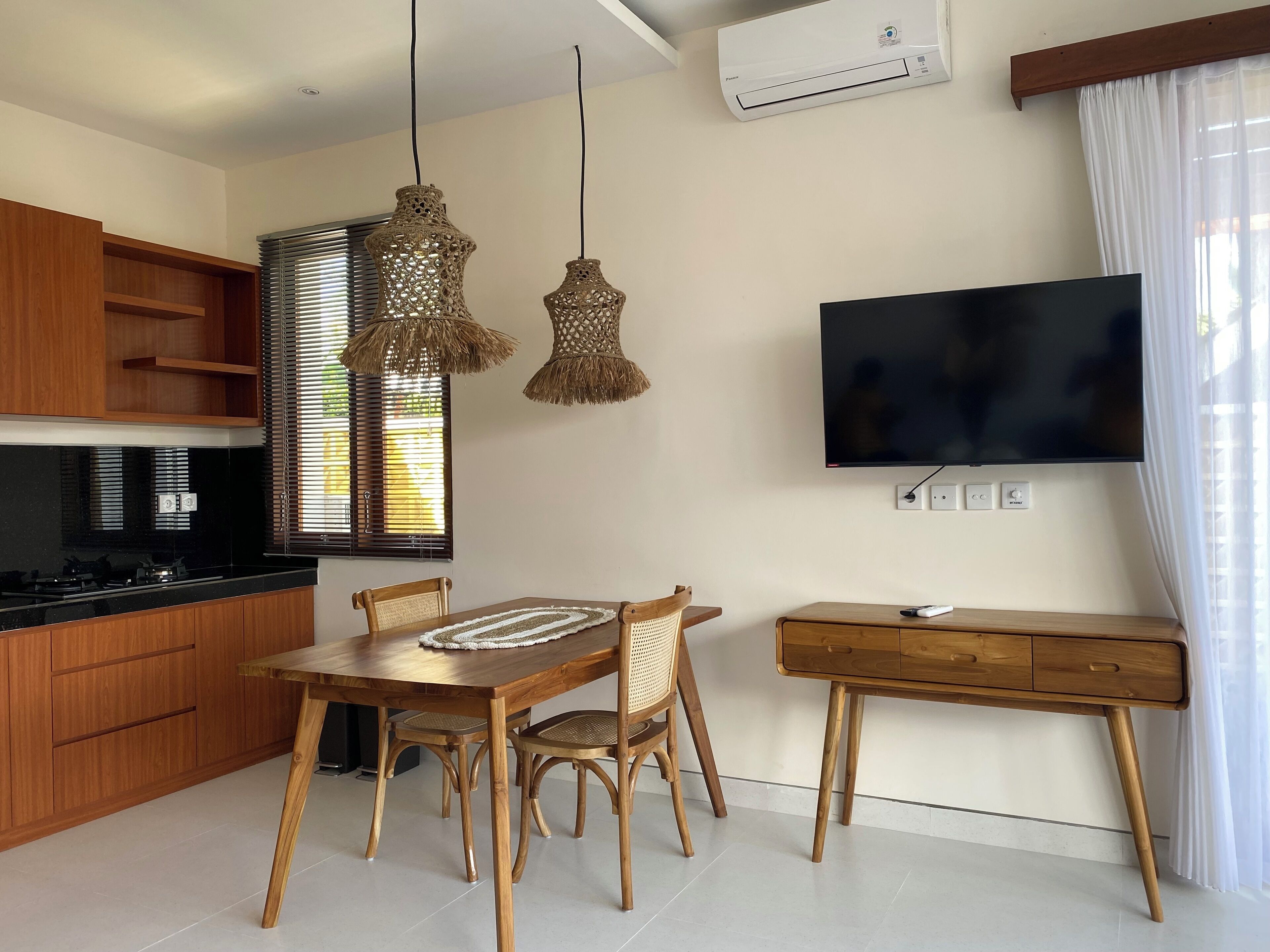 Cozinha moderna com armários de madeira, bancada preta e área de jantar com mesa e cadeiras de madeira. Há uma TV montada na parede e um ar condicionado acima dela.