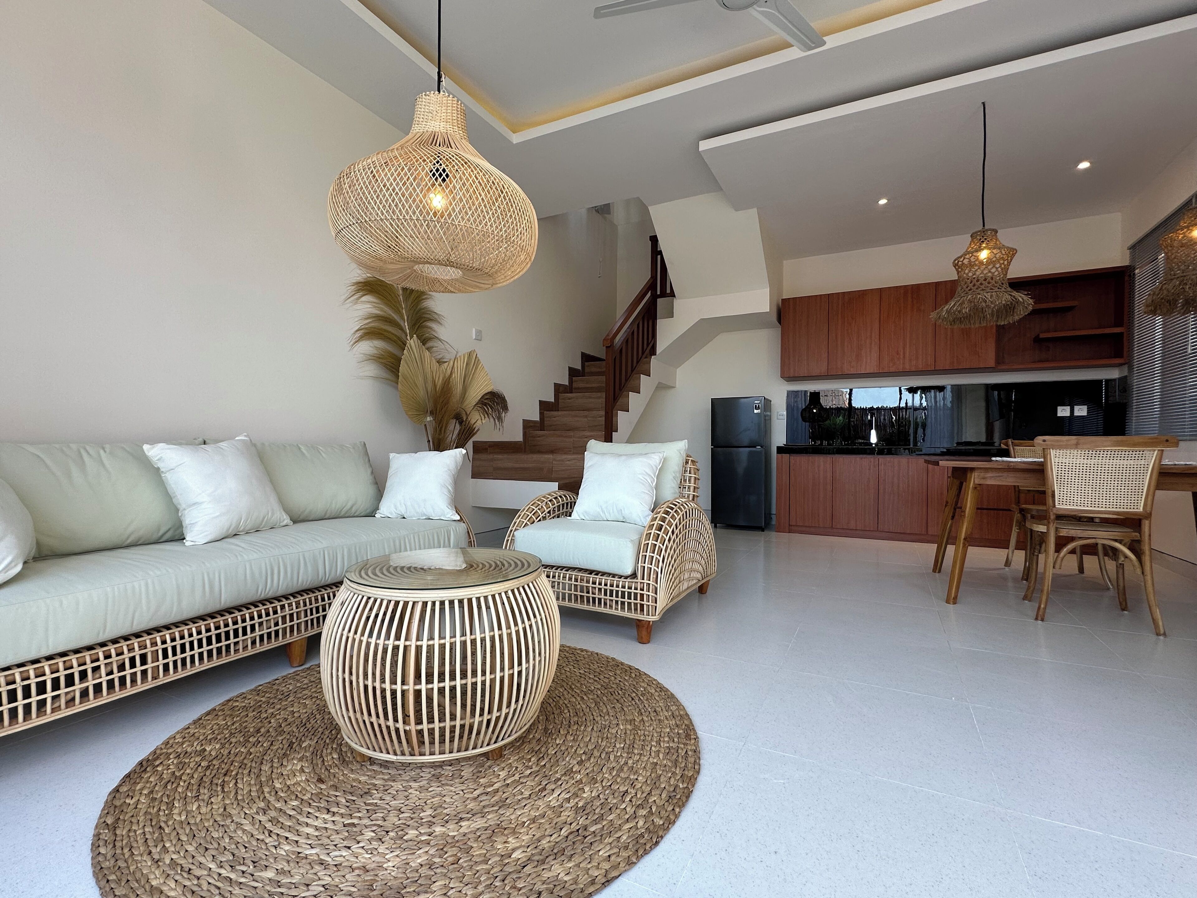 A modern living room with a sofa, coffee table, and wicker chairs. A kitchen with wooden cabinets and a refrigerator is visible in the background.