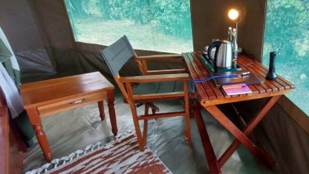 Ein Holztisch mit einem Laptop, einer Lampe und einem Notizbuch in einem zeltartigen Raum mit Blick auf Bäume.