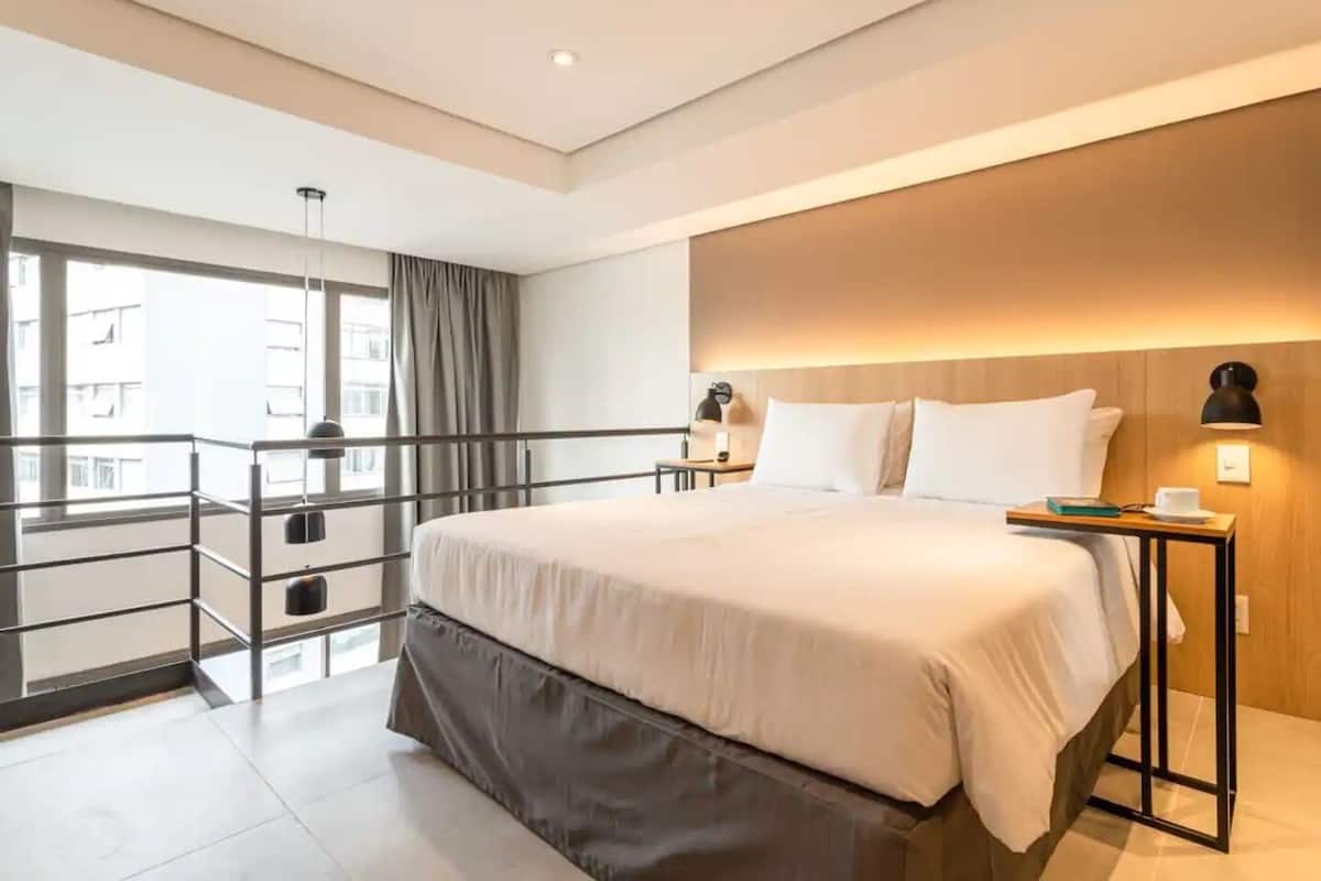 Une chambre d’hôtel moderne dotée d’un grand lit, d’une table de chevet, d’un bureau, d’un balcon avec une rambarde et offrant une vue sur les bâtiments environnants.