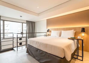 A modern hotel room with a large bed, a nightstand, a desk, a balcony with a railing, and a view of buildings outside.