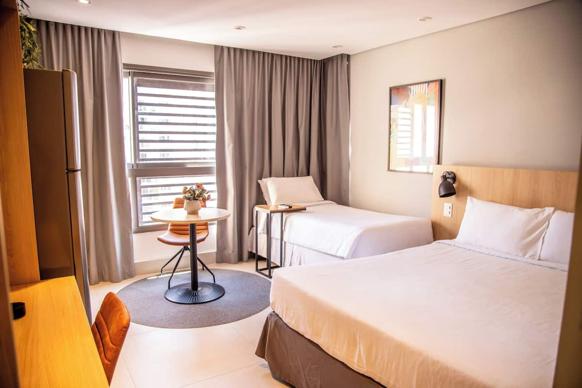 Une chambre d’hôtel avec un lit, un bureau, une chaise, une petite table avec une plante et une fenêtre avec des rideaux.