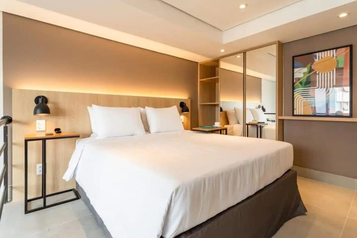 Moderni hotellihuone, jossa on suuri sänky, yöpöydät, vaatekaappi ja taulu seinällä.