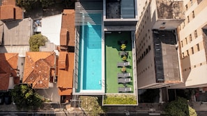 An aerial view of a hotel with a rooftop pool, multiple balconies, and a green lawn area with sun loungers.
