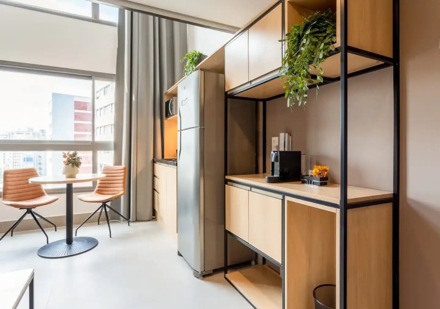 A modern kitchenette with a refrigerator, coffee maker, and a small plant. There is a dining area with two chairs and a table, and a window with a view of buildings.