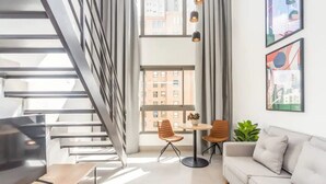 A modern living room with a staircase, a sofa, two chairs, a small table, and a large window with city views. - Cozzy Suites Paraiso Hotel (São Paulo)