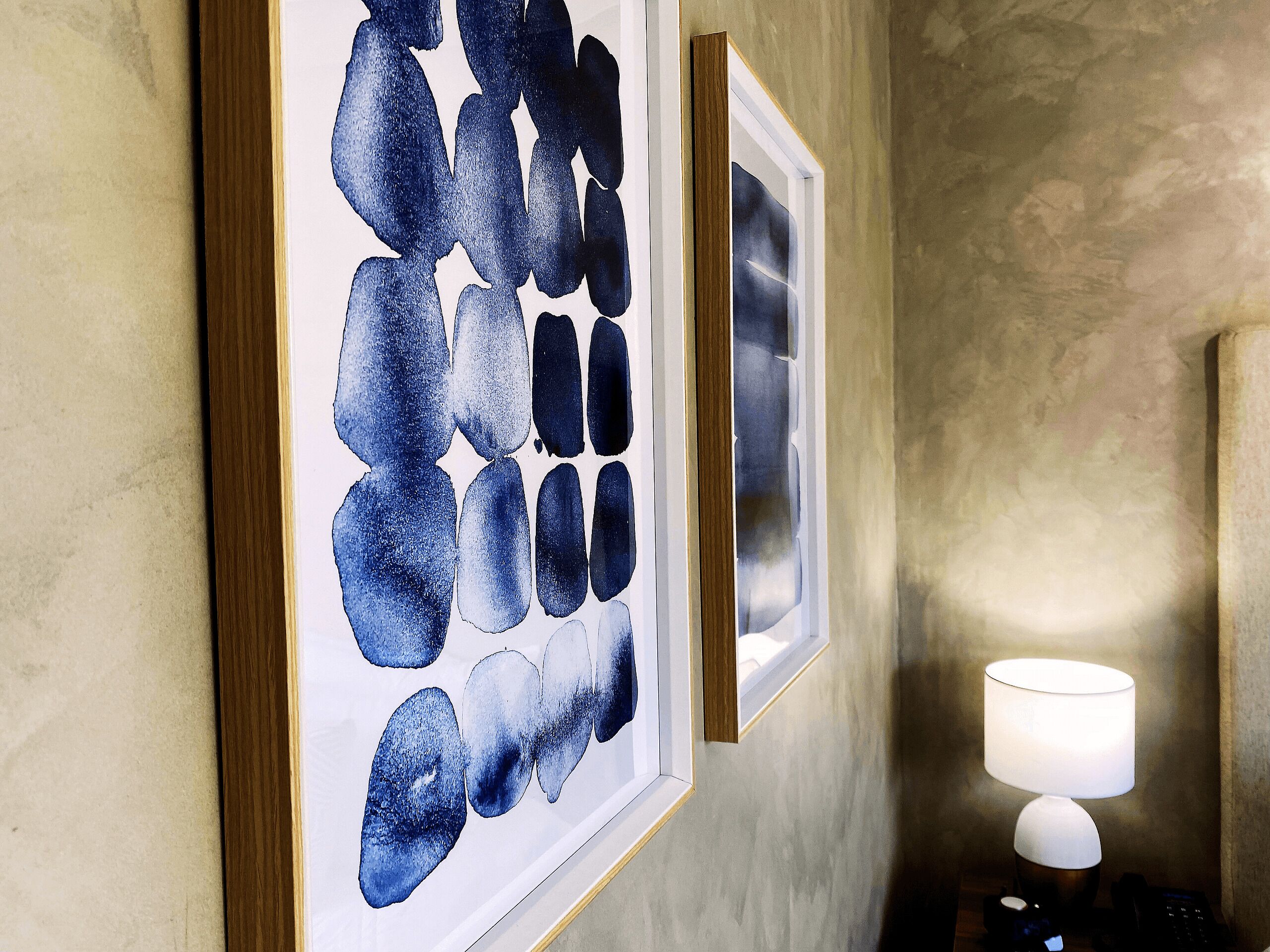 Een gang met abstracte kunstwerken in lijsten aan de muur en een lamp op een tafel.