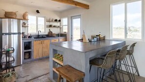 A modern kitchen with a central island, wooden beams, and a large window. - Casa Flamingo | Cozy Cabin with Views | 5 Acres (Yucca Valley)
