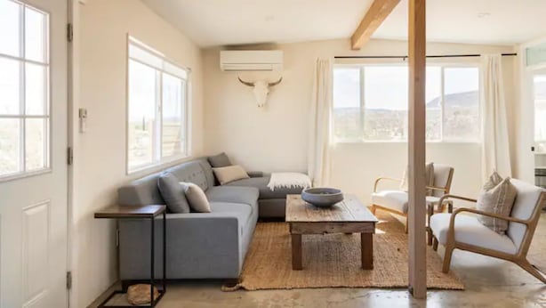 A modern living room with a grey sofa, a wooden coffee table, and a bull skull wall decoration. - Casa Flamingo | Cozy Cabin with Views | 5 Acres (Yucca Valley)