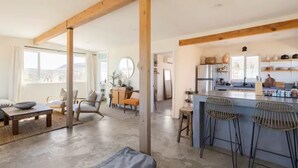 A spacious living area with a large window, a kitchen with bar stools, and a dining area with a table and chairs. - Casa Flamingo | Cozy Cabin with Views | 5 Acres (Yucca Valley)