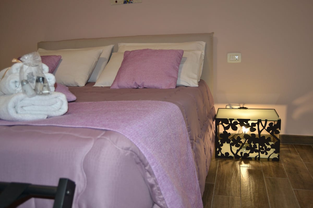 Ein ordentlich gemachtes Bett mit einer rosa Bettdecke und Kissen, ein Nachttisch mit einer dekorativen Lampe und ein gefaltetes Handtuch auf dem Bett.
