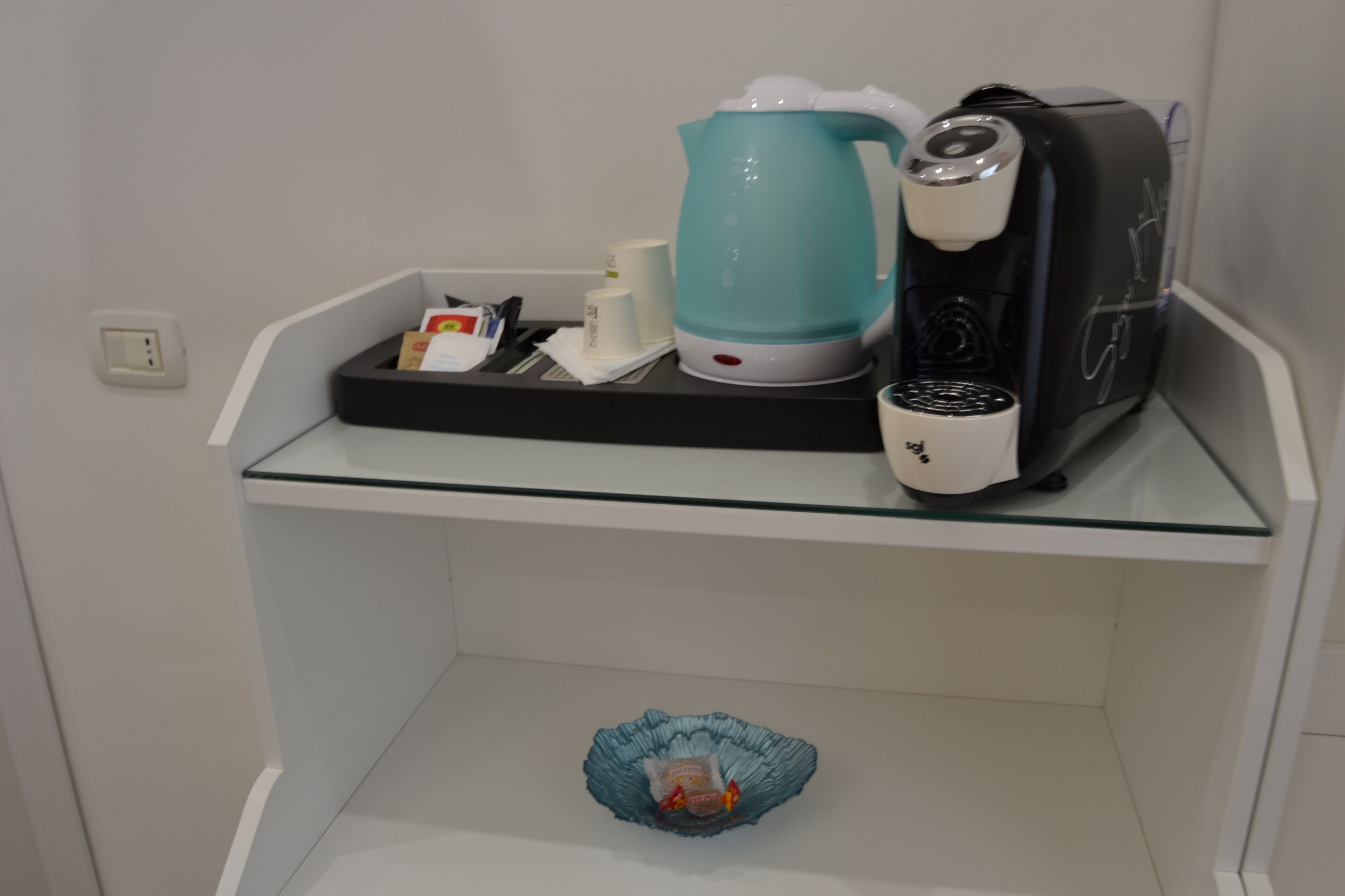 Una zona de cocina con cafetera, hervidor y microondas sobre una repisa de vidrio.