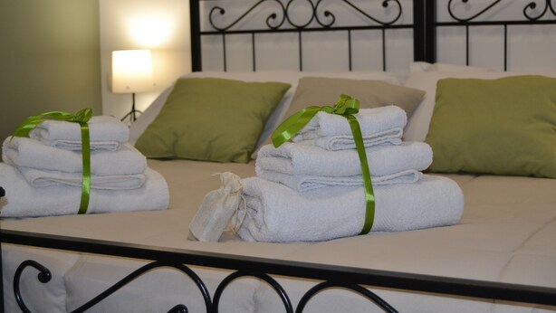 Una cama bien hecha con almohadillas verdes y toallas dobladas, una lámpara de noche y un cabecero de metal.