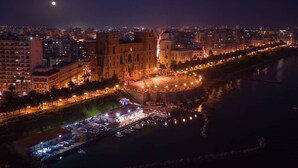Un paisaje urbano nocturno con un edificio histórico destacado, un paseo marítimo y una plaza circular.