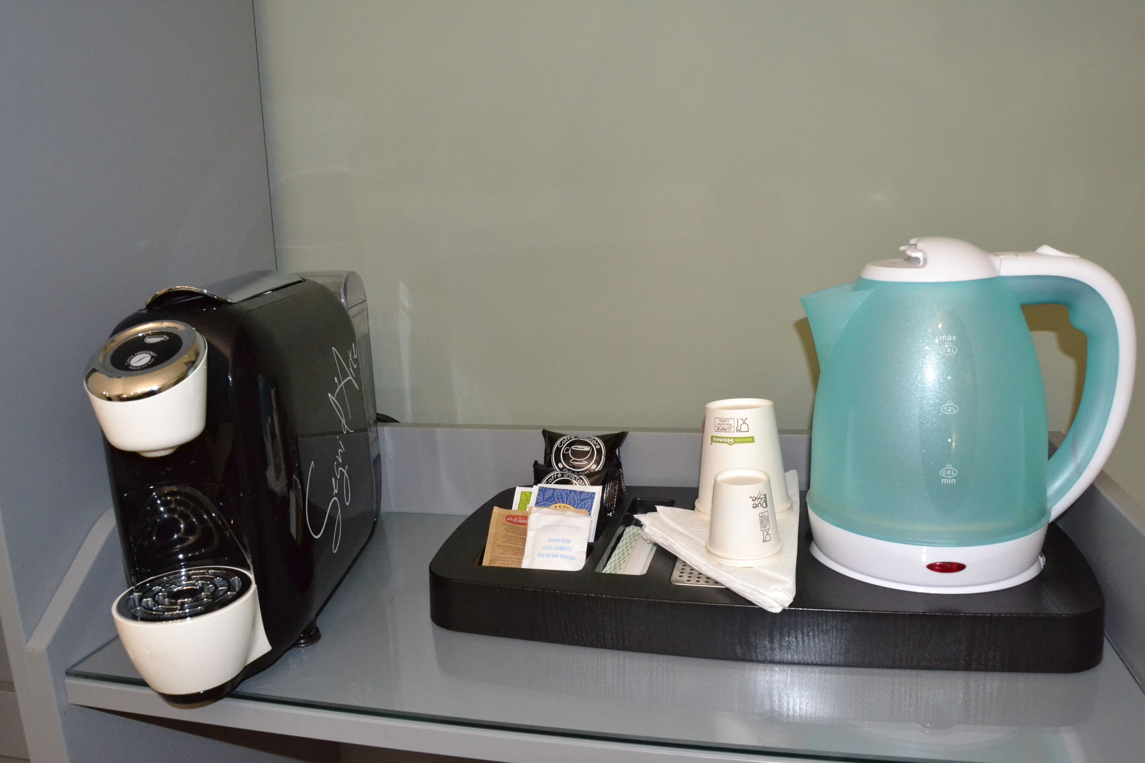 A coffee maker, a kettle, and a tray with coffee pods and a cup on a hotel minibar shelf.