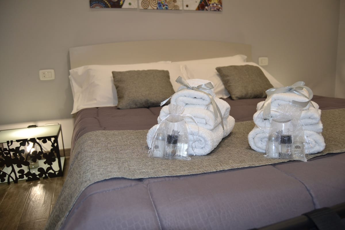 Ein ordentlich gemachtes Bett mit aufgerollten Handtüchern und Toilettenartikeln, ein Nachttisch mit Lampe und eine Wand mit einem Gemälde.