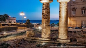 Columnas de piedra antiguas en un sitio histórico, con vista al mar y edificios modernos al fondo.