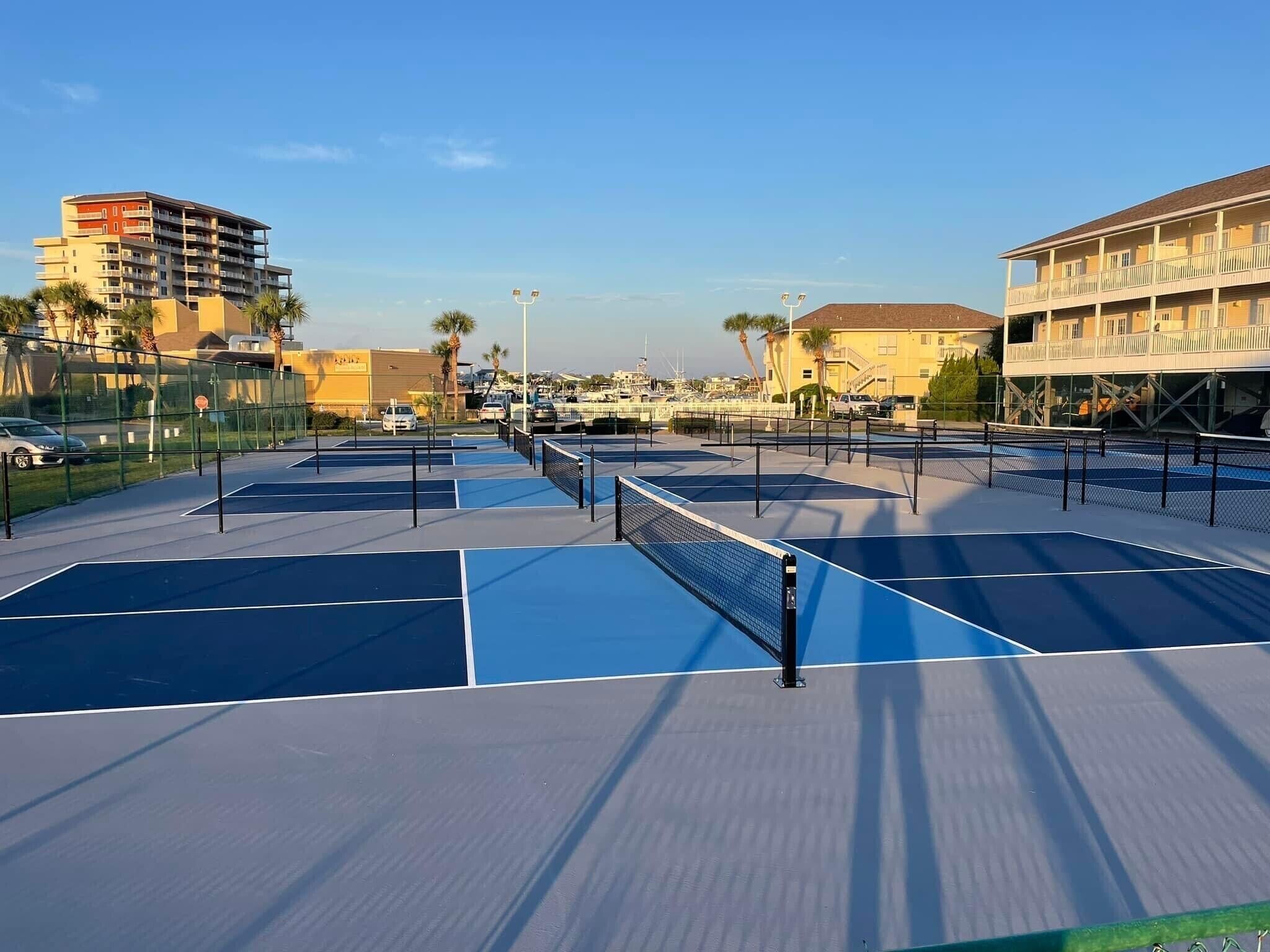 Un court de tennis extérieur comprenant plusieurs terrains, entouré d’une clôture, avec des bâtiments en arrière-plan.