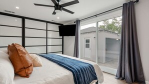 A modern bedroom with a bed, a ceiling fan, a TV, and a sliding glass door to the outside. - Luxury Hangout | Private Spa + Game Room + Arcade (Long Beach)