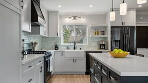 A modern kitchen with white cabinets, a stainless steel refrigerator, and a large island with a bowl of fruit. - Luxury Hangout | Private Spa + Game Room + Arcade (Long Beach)
