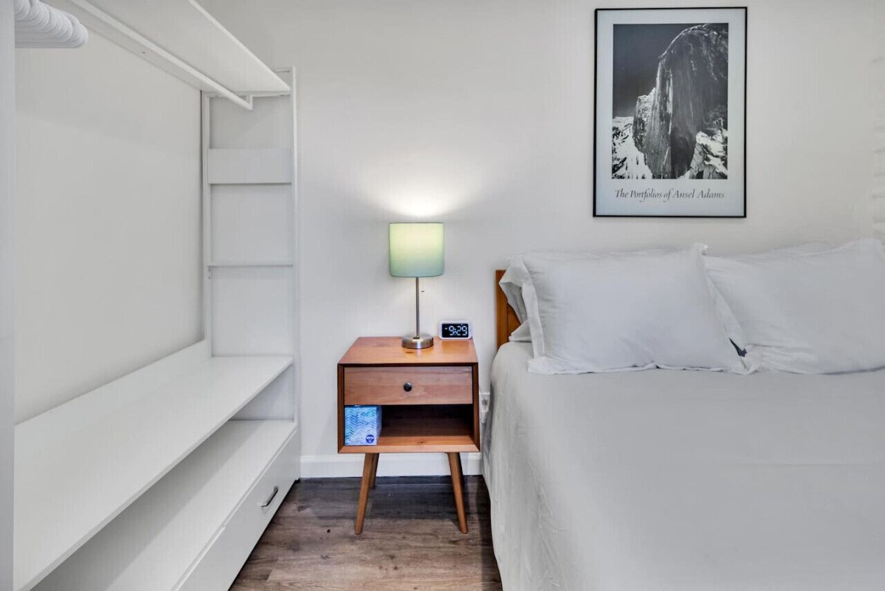 Een slaapkamer met een bed, een nachtkastje met een lamp, een lijst met een foto aan de muur en een houten plank.