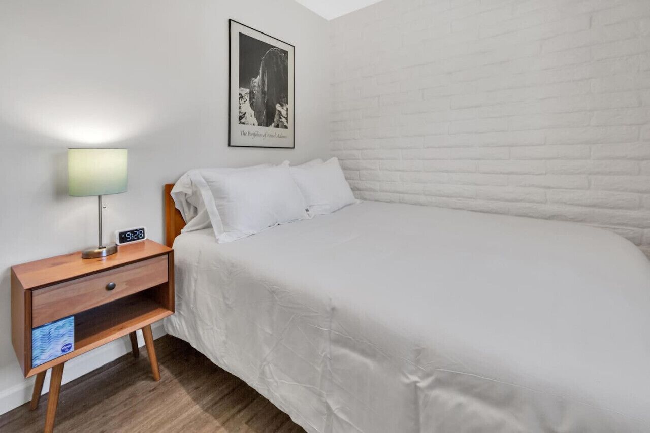 A bedroom with a white brick wall, a bed with white bedding, a wooden nightstand with a lamp, and a framed picture on the wall.