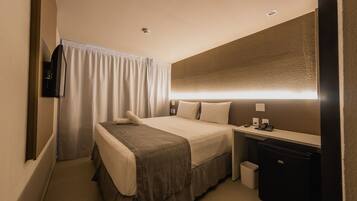 Quarto de hotel com cama, mesa de cabeceira, abajur e um cesto de lixo pequeno.