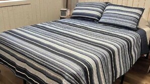 A bed with a striped bedspread and pillows, a bedside lamp, a window with blinds, and a wooden floor. - Cozy Tiny Home Cabin Near Lake Greeson & Kirby Landing – Pet-Friendly! (Kirby)