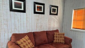 A room with a brown sofa, three framed pictures on the wall, and a window with blinds. - Cozy Tiny Home Cabin Near Lake Greeson & Kirby Landing – Pet-Friendly! (Kirby)