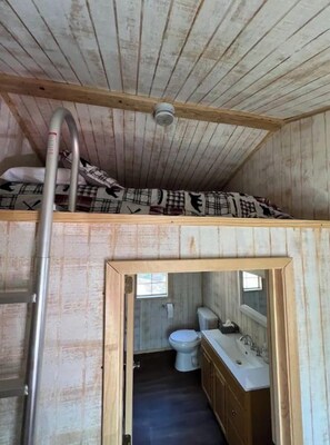 A small, rustic cabin with a loft bed, a wooden ladder, a bathroom with a toilet and sink, and a window for natural light. - Spacious Double Loft Cabin Near Lake Greeson Pet-Friendly– Sleeps 8 (Kirby)