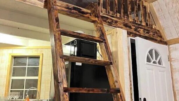 Een houten ladder die leidt naar een hoger gelegen gedeelte, met een zwarte koelkast en een witte deur.