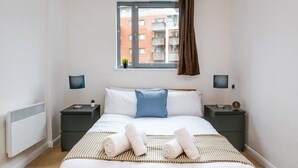 Une chambre à coucher bien rangée, avec un grand lit, deux tables de chevet, une fenêtre avec des rideaux et une vue sur les bâtiments à l’extérieur.